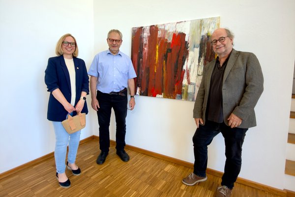Drei Personen posieren vor einem Gemälde für ein Foto. Links steht eine Frau mit dunkelblauem Blazer, in der Mitte ein Mann mit einem hellblauen Hemd und rechts ein Mann mit einem grauen Sakko.
