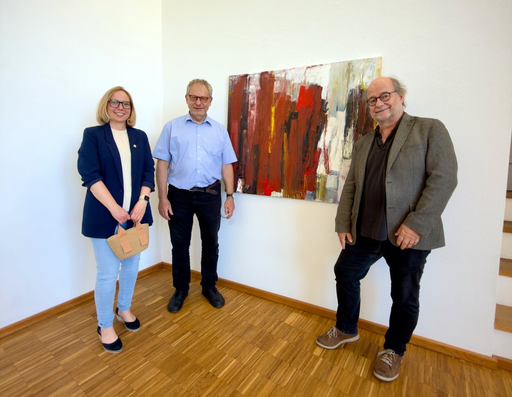 Drei Personen posieren vor einem Gemälde für ein Foto. Links steht eine Frau mit dunkelblauem Blazer, in der Mitte ein Mann mit einem hellblauen Hemd und rechts ein Mann mit einem grauen Sakko.