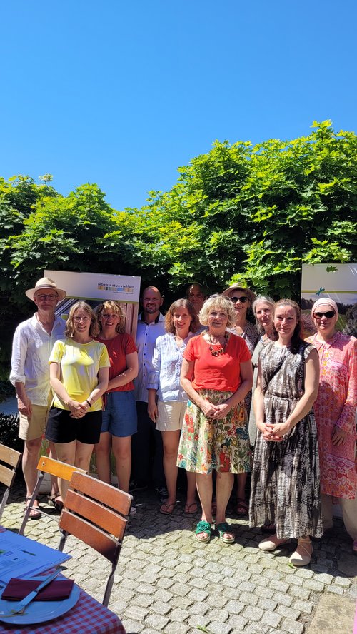 Mehrere Personen posieren als Gruppe für ein Foto. Sie sind in einem Biergarten. Die Sonne scheint. 