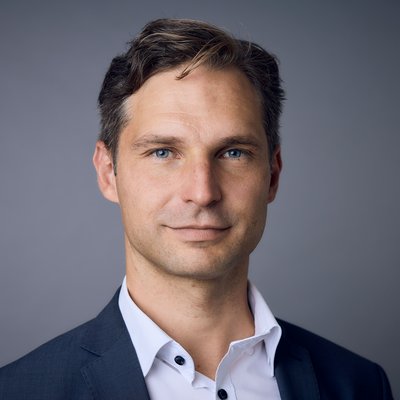 Ein Mann schaut in die Kamera. Er trägt ein dunkles Sakko und ein weißes Hemd. Seine Haare sind dunkelblond und kurz.