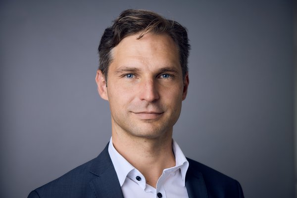 Ein Mann schaut in die Kamera. Er trägt ein dunkles Sakko und ein weißes Hemd. Seine Haare sind dunkelblond und kurz.