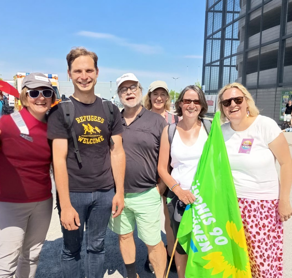 Sechs Personen sehen in der Sonne. Drei Personen tragen eine Mütze. Eine Person trägt eine grüne Fahne.