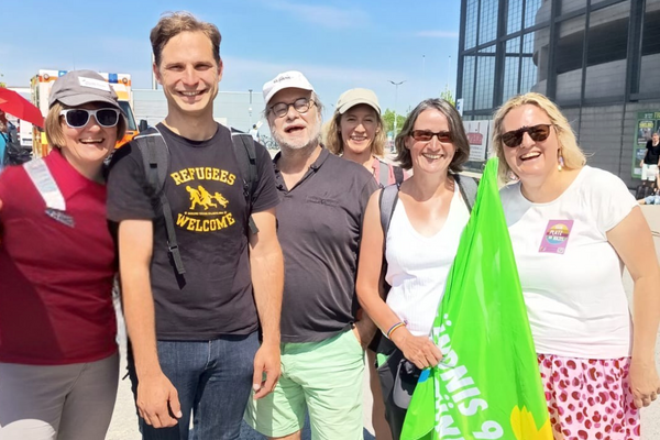 Sechs Personen sehen in der Sonne. Drei Personen tragen eine Mütze. Eine Person trägt eine grüne Fahne.