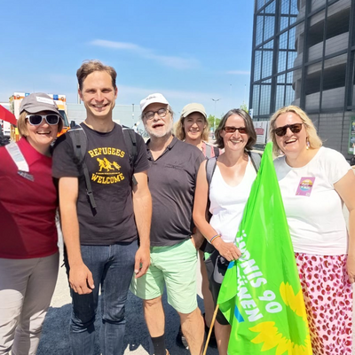 Sechs Personen sehen in der Sonne. Drei Personen tragen eine Mütze. Eine Person trägt eine grüne Fahne.