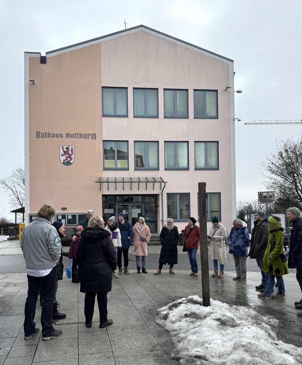 Rundgang mit der Wählergemeinschaft LebensWert Hutthurm Etwa 15 Personen stehen vor dem Rathaus in Hutthurm. Sie hören einem Vortrag zu.