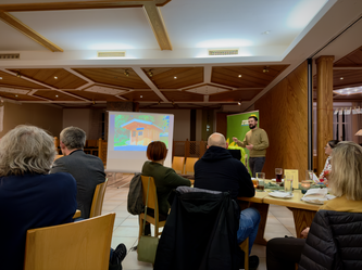 Landkreis-Grüne besuchen Diskussionsveranstaltung in Böhmzwiesel Ein Mann hält einen Vortrag vor einer Gruppe in einem Gastraum mit Holzvertäfelung. Auf der Leinwand hinter ihm ist eine Präsentation mit einem Haus zu sehen. Die Zuhörenden sitzen an Tischen mit Getränken und Unterlagen, einige machen sich Notizen.