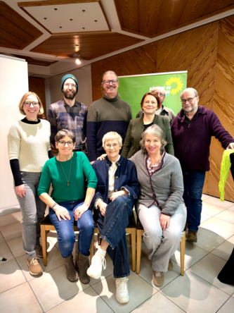 Landkreis-Grüne besuchen Diskussionsveranstaltung in Böhmzwiesel Neun Personen, drei sitzend und sechs stehend, lächeln in die Kamera bei einer Veranstaltung mit grünem Banner im Hintergrund.