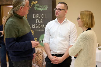Landkreis-Grüne besuchen Diskussionsveranstaltung in Böhmzwiesel Drei Personen stehen vor einem Banner der Bio-Bäckerei Wagner und diskutieren, ein Mann hält einen Stift in der Hand. Im Hintergrund hängt ein Banner mit dem Logo der Bio-Bäckerei Wagner und den Worten EHRLICH. FAIR. REGIONAL! sowie der Webadresse www.wagner.bio.