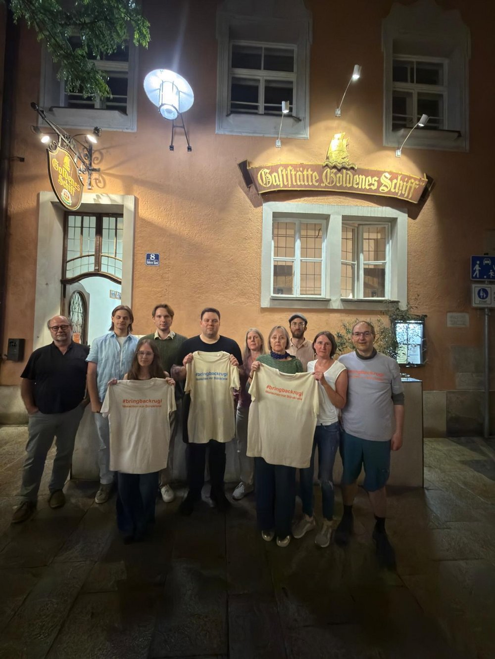 Zehn Personen halten vor dem Gasthaus "Goldenes Schiff" in Passau mehrere T-Shirts mit der Aufschrift "#bringbackrugi"