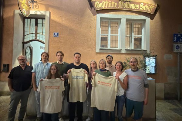 Zehn Personen halten vor dem Gasthaus "Goldenes Schiff" in Passau mehrere T-Shirts mit der Aufschrift "#bringbackrugi"