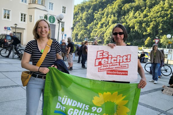 Zwei Frauen halten eine Fahne und ein Plakat. Auf dem Plakat steht "Es reicht. Das Leben bezahlbar machen. Niederbayern". Die Fahne ist grün und darauf steht "Bündnis 90 / Die Grünen".