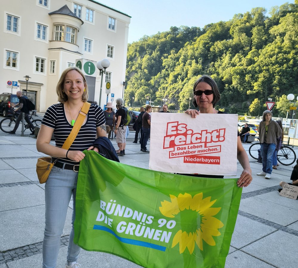 Zwei Frauen halten eine Fahne und ein Plakat. Auf dem Plakat steht "Es reicht. Das Leben bezahlbar machen. Niederbayern". Die Fahne ist grün und darauf steht "Bündnis 90 / Die Grünen".